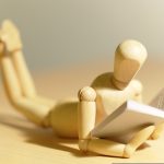 Wooden mannequin figure lies on stomach reading a book on a table surface.