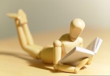 Ask the trainer: “What are some exercises I could do while studying or sitting in class?” Wooden mannequin figure lies on stomach reading a book on a table surface.
