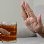 Are you sober curious? Explore the benefits of drinking less and living better Close-up of a person holding up their hand to decline a glass of amber-colored liquid being offered by someone else.