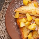 Plated vegan French toast topped with warm fruit pieces and powdered sugar on rustic wooden table.