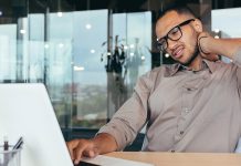 Ask the trainer: “What exercises can help relieve neck pain from sitting at my desk too long?” A man rubbing his neck while using laptop, showing neck pain from desk work.