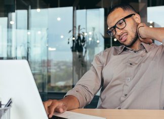Ask the trainer: “What exercises can help relieve neck pain from sitting at my desk too long?” A man rubbing his neck while using laptop, showing neck pain from desk work.