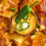 Plate of nachos topped with melted cheese, bacon bits, and sliced jalapeños, served with a bowl of cheese dip garnished with peppers.