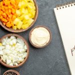 Top-down view of wooden bowls with chopped vegetables, eggs, and dressing, next to a notepad labeled ‘5 plant-based recipes.’