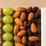 Neatly arranged snack ingredients including grains, grapes, almonds, raisins, and protein bar on neutral background.