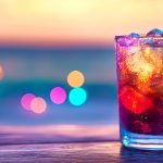 Colorful sparkling drink with ice, lime, and pine garnish against a blurred festive background.