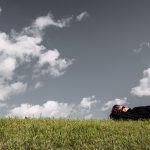 Person lies on grassy hill under cloudy sky, embracing stillness and mindfulness outdoors.