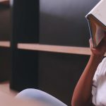 Student overwhelmed, sitting with a book covering their face in a study area.