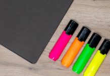 A black notebook and four colorful highlighters lie on a wooden desk, ready for studying.