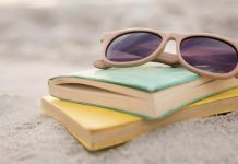 Two books stacked on sandy beach with sunglasses resting on top, evoking summer reading relaxation.