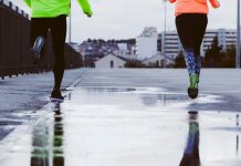 Two people running on a wet road in urban surroundings, wearing bright jackets on a cloudy day.
