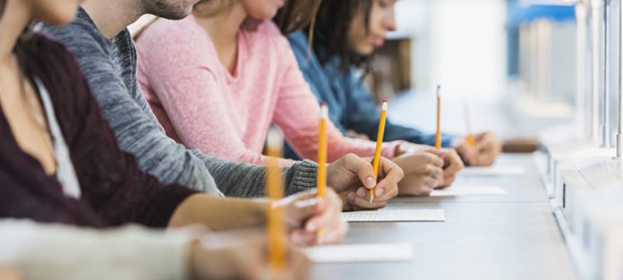 Cropped view of a multiracial group of young men and women sitting in a row at a table, writing with pencils on paper. They are taking a test or filling out an application. Focus is on the hand of the young man in the middle in the gray shirt.